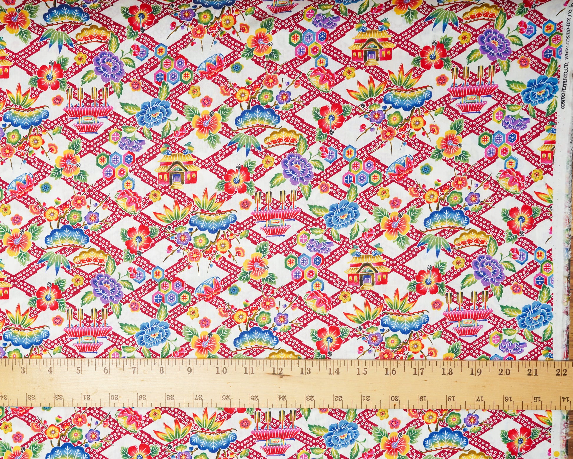 Colorful fabric pattern with geometric design and floral elements on a white background, measured with a ruler at the bottom.